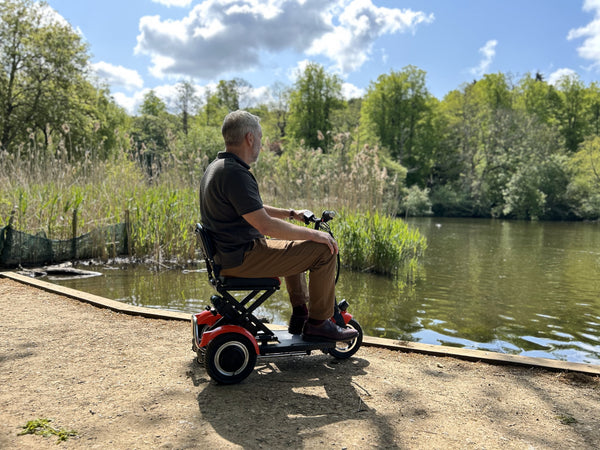 A man riding a folding mobility scooter near a serene lake, enjoying the scenic beauty of nature.