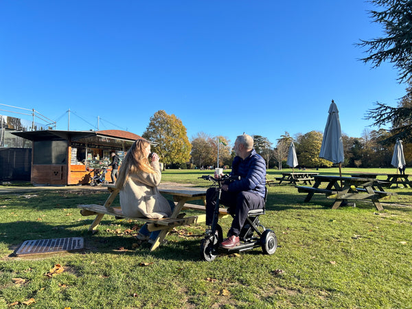 A woman and a man on a folding mobility scooter sitting at a picnic table enjoying a leisurely outdoor meal together.