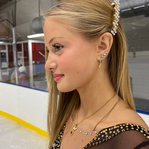 figure skater standing on ice wearing gold ice skating earrings in a brown sparkly skating dress