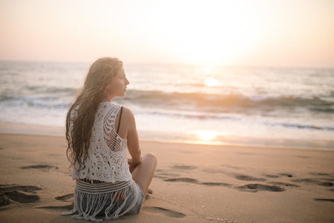femme assise sur le sable, face à la mer et au coucher de soleil portant un haut dentelle à frange