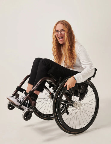 Ella, a white woman with long curly blonde hair. She is wear a white shirt, black trousers and is using a manual wheelchair