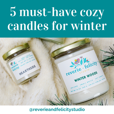 A Hearthside scented soy wax candle is nestled into a white faux fur blanket next to a larger Winter Woods scented candle in this image from an article frm 5 must-have cozy candles for winter from Reverie + Felicity Studio