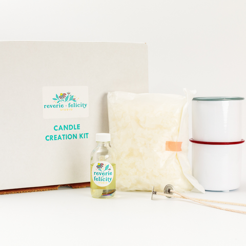 An arrangement of candle-making supplies sits in front of a white background. From left to right is a white box, a glassine bag of wax flakes, a clear glass bottle of fragrance oil, a pair of wicks and two white enamel candle vessels. Reverie + Felicity Studio DIY candle making kit candle creation kit make your soy candles