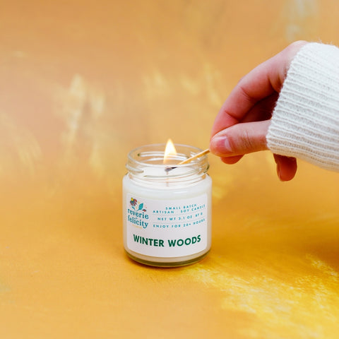 A light-skinned hand hugged by a cream-colored sweater holds a lit match to the wick of a candle. The candle is made of white soy wax poured in a clear glass jar and sits against a mustard yellow backdrop. Winter Woods soy candle from Reverie + Felicity Studio evergreen balsam fir