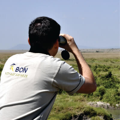 male guide putting on a white shirt with writtings of Bon Voyage Kenya Safaris looking through a binocular at daytime in Masai Mara