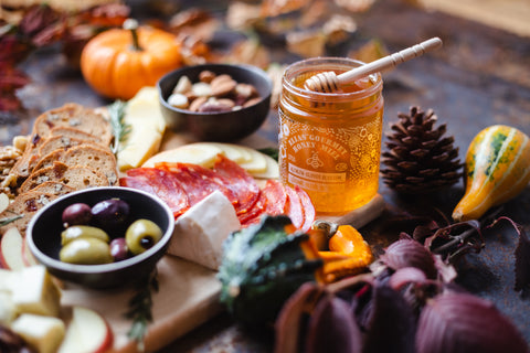 Honey dipper dipped into Raspberry Gourmet Honey Jar, Charcuterie board in the background