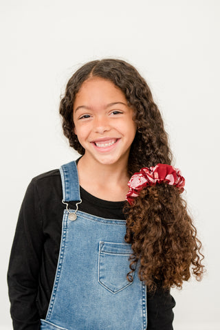 Young girl with curly hair in crepe silk scrunchie. Girl is wearing black shirt, and denim overalls.