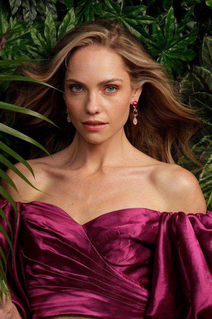 Woman in a pink off the shoulder top stands in front of a green leafy wall. She's wearing Kelly Woodcroft mermaid drop stone earrings with beautiful handcrafted details that are made in her Brisbane studio.