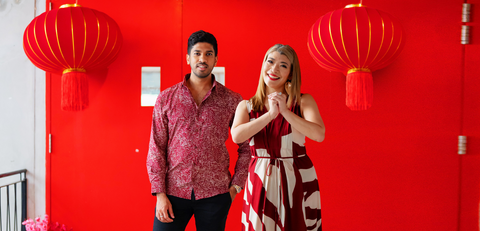 a man and woman in vibrant red colors posing in front of a red door with chinese new year elements as decoration as they celebrate chinese new year in authentic batik
