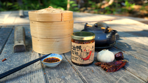 A steam basket, a jar of Chinese Chili Oil, a sauce dish with some chili oil, a pair of chopsticks are in the foreground. In the background, there are a star anise, a bulb of garlic, and a Chinese teapot.