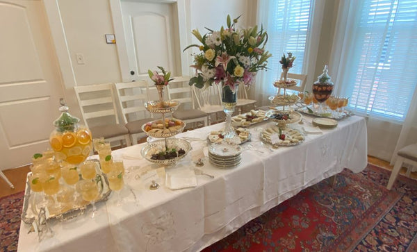 A table with white linens, silver trays, and clear carafes of tea ready for a tea party.