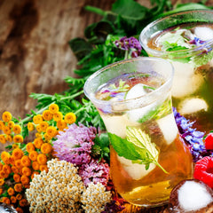 Glass or iced herbal tea surrounded by vibrantly colored herbs and flours of many types.