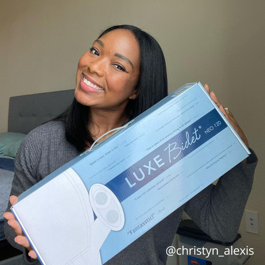 Smiling woman happily holding up her brand new LUXE Bidet box. Text in the corner shows the post is from @christyn_alexis on Instagram