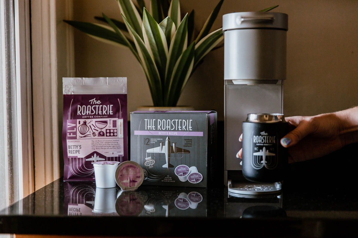 A box of Betty's Recipe flavored single serve cups sits next to a k-cup style brewer, with a person's hand grabbing a mug of coffee.