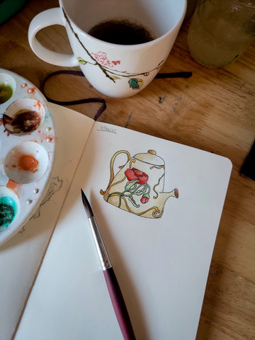 an open sketchbook with a drawing and watercolor painting in it sitting next to a tea mug with some tea in it.