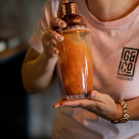 Women shaking copper topped glass cocktail shaker with red brown coloured liquid to make a cold brew negroni