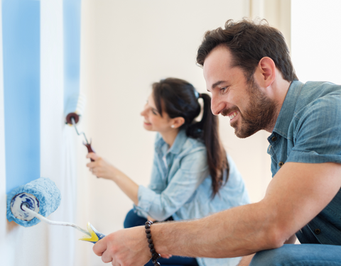 Egy férfi és egy nő közösen festik a falat, a férfi egy hosszú nyelű hengerrel, a nő egy kisebb ecsettel dolgozik, mindketten összpontosítanak a feladatra boldog kifejezéssel.