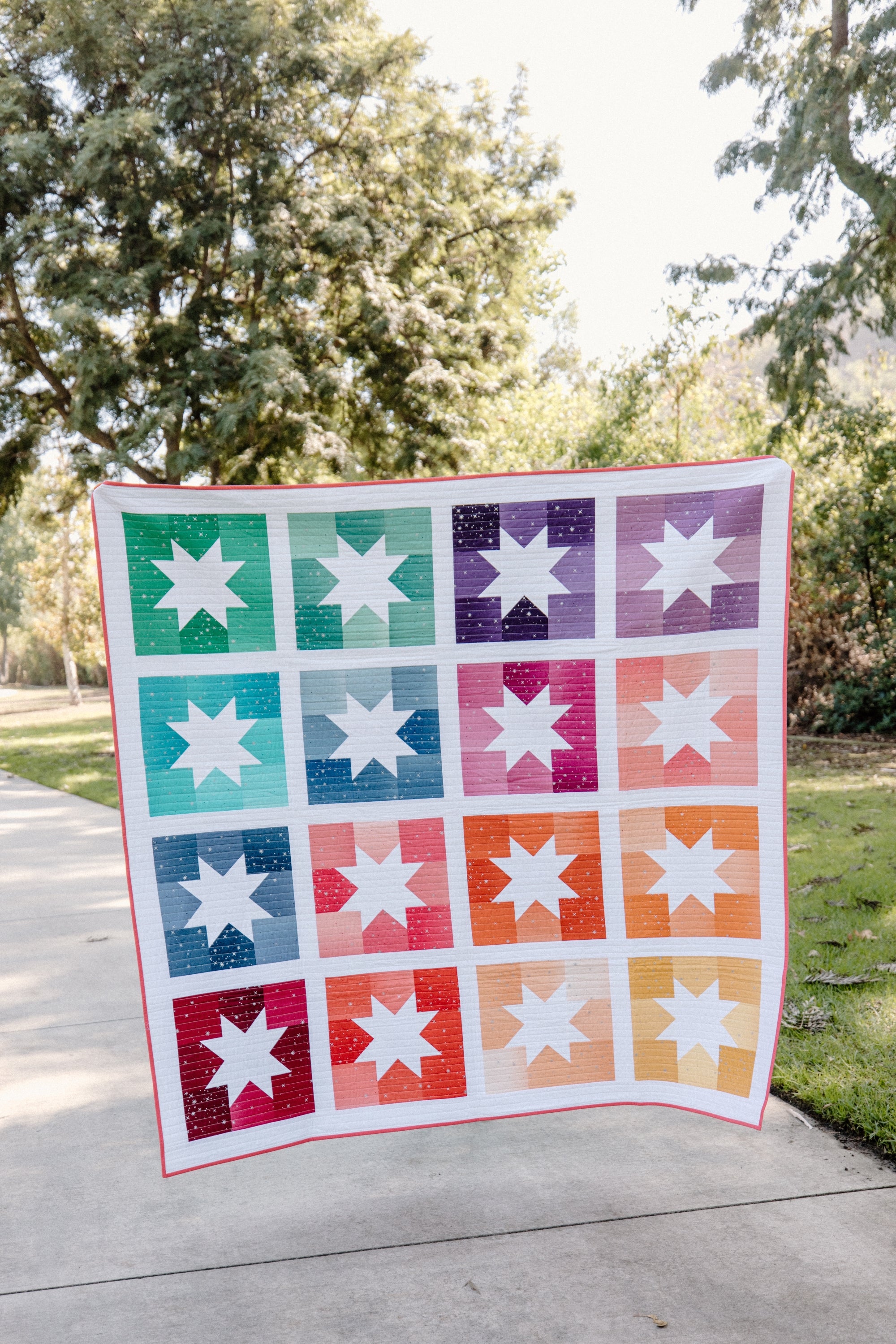 Joyful Stars quilt, Cotton and Joy’s Pattern of the Month, held outdoors on a sunny day. The quilt features sawtooth star blocks in vibrant ombre shades of green, blue, purple, pink, orange, and yellow, set against a white background.