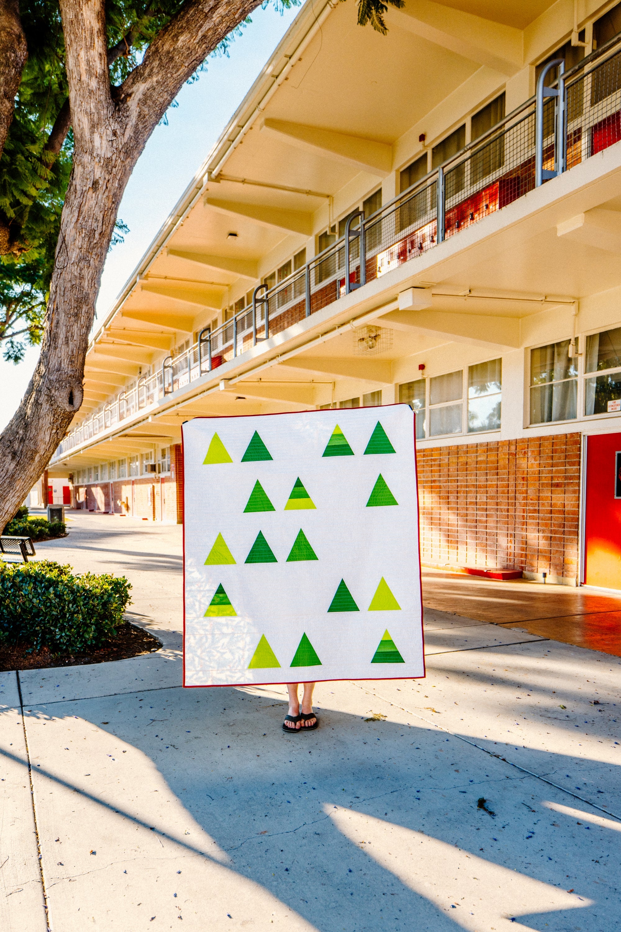 Tree Farm quilt pattern by Cotton and Joy, featuring green triangle trees on a white background, held outdoors in front of a mid-century modern building with sunlight and shadows.