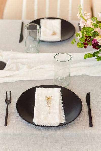 Table decorations including black plates, black cutlery, and a white table runner on gray linens create a dazzling tablescape - sold by Bluum Maison.