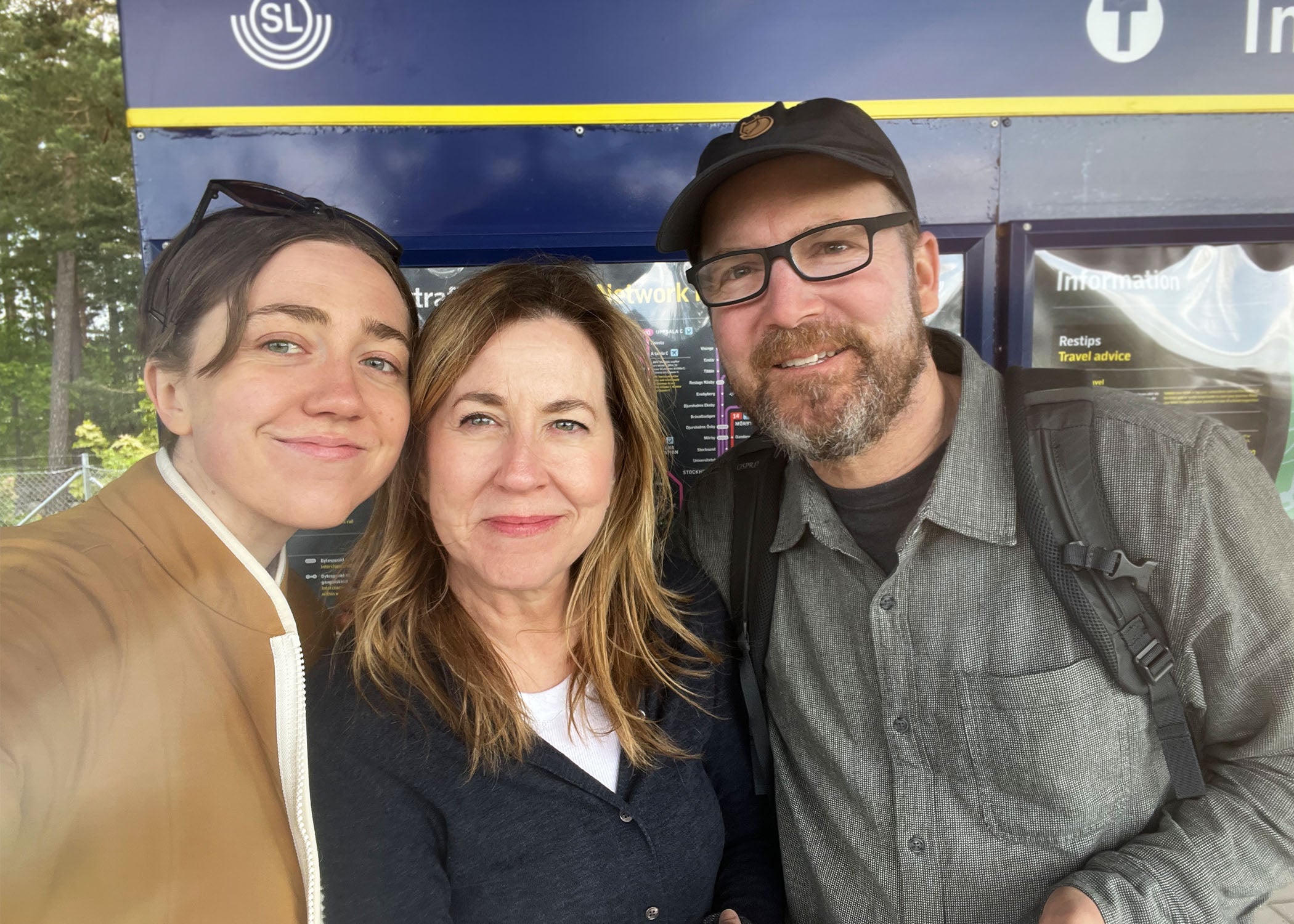 Three people on the Woodland Mod travel team smile in front of a train stop in Sweden