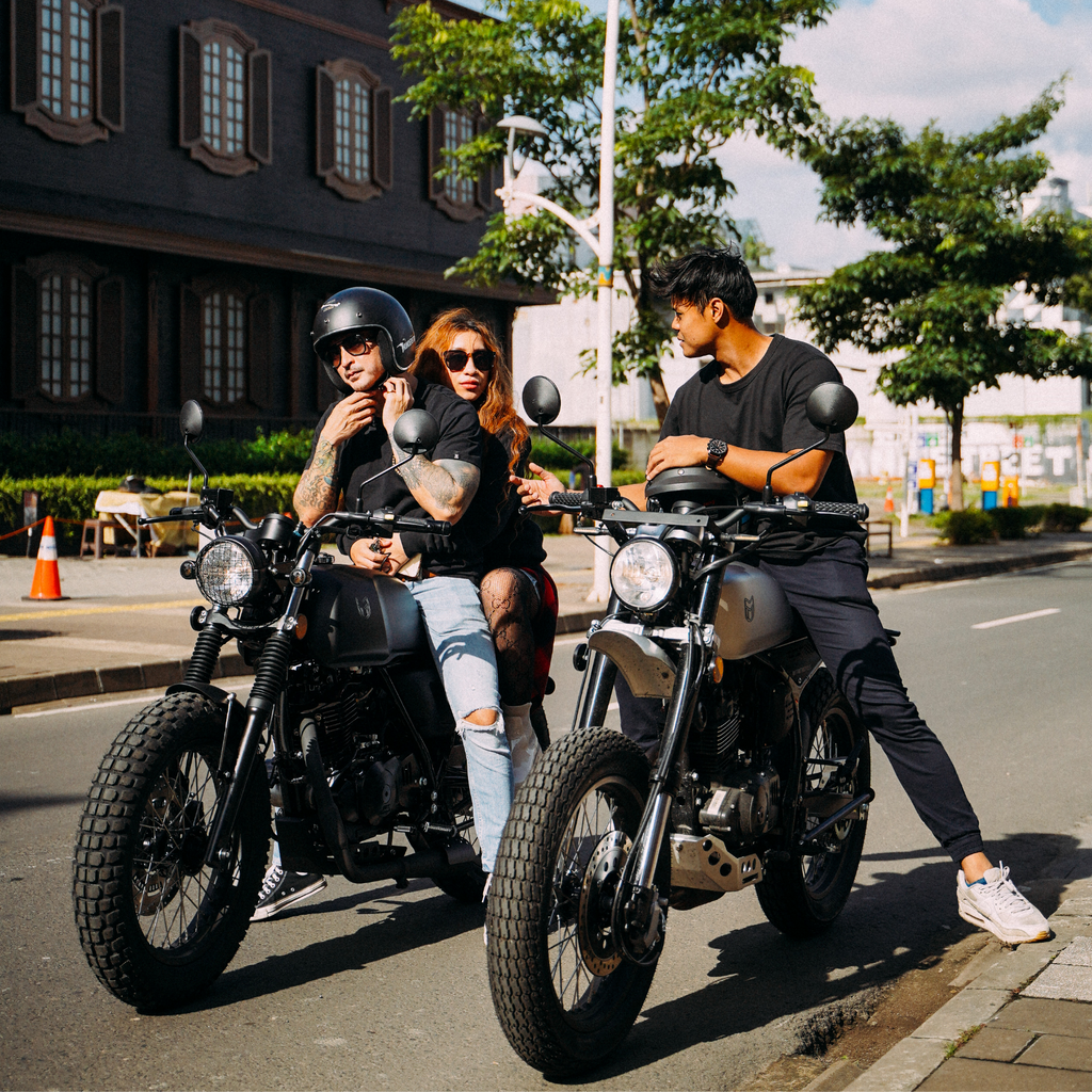 A sunny day with 3 people riding mutt motorcycles wearing sunglasses. One is talking to the people on the motorcycles next to him as the other rider adjusts their helmet.