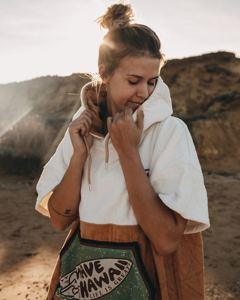 Eine Frau im Poncho steht am Strand in der Sonne und kuschelt sich in die Kapuze ein.