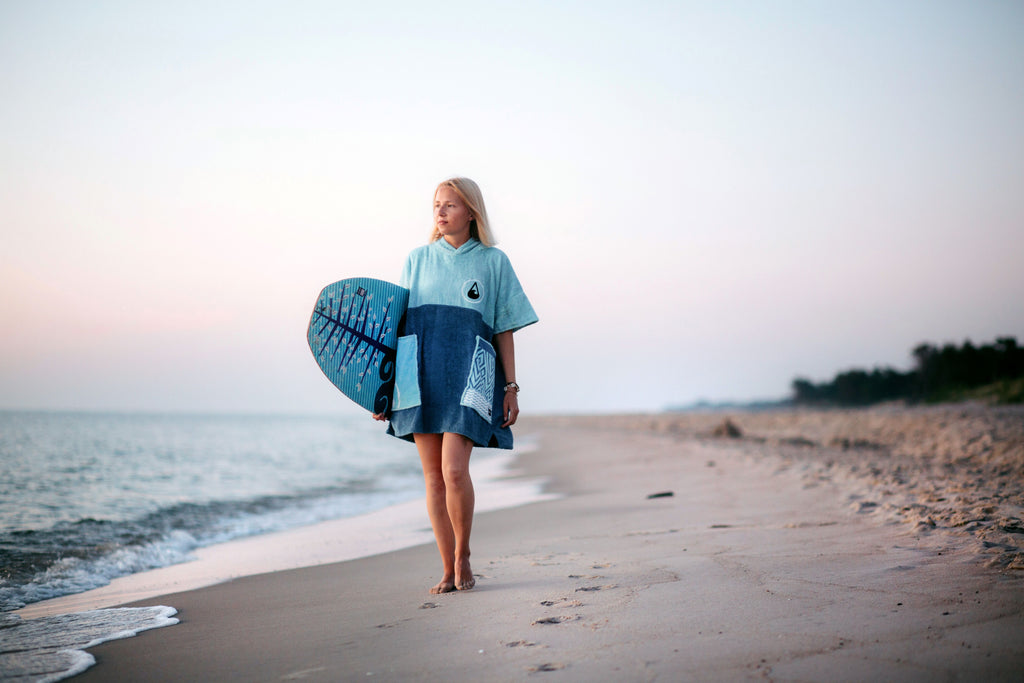 Eine Frau im blauen Poncho läuft am Strand entlang mit einem Surfboard unter dem Arm.