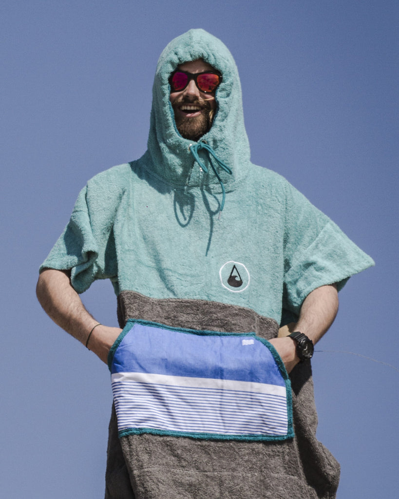 Ein lachender Mann im Poncho mit Händen in der Bauchtasche und etwas zugezoogener Kapuze steht in der Sonne.