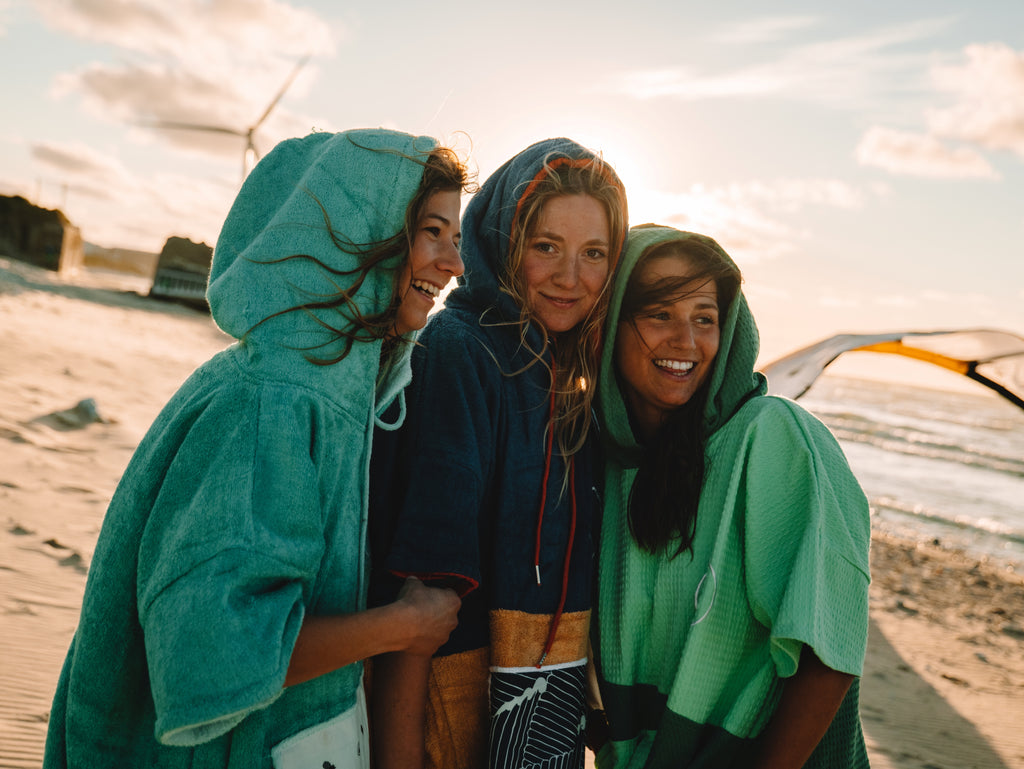 Drei Frauen im Poncho stehen nebeinander am Strand mit der Sonne im Hintergrund.