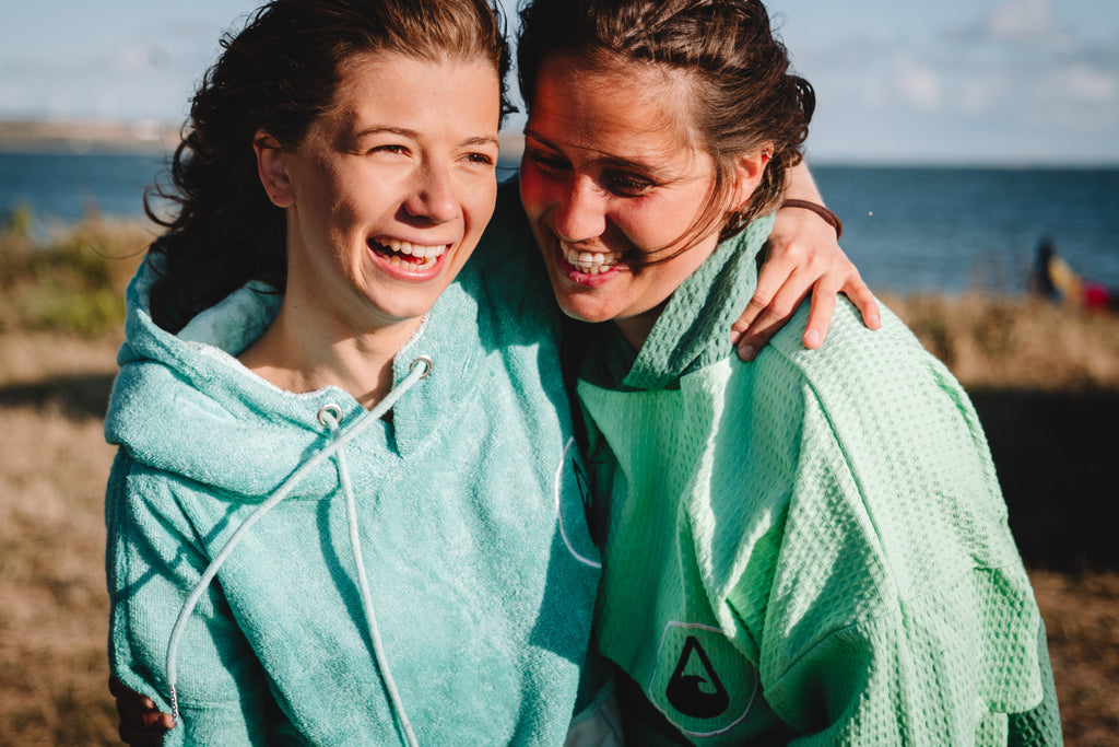 Zwei lachende Frauen laufen Arm in Arm mit ihren Ponchos bekleidet über den Strand.