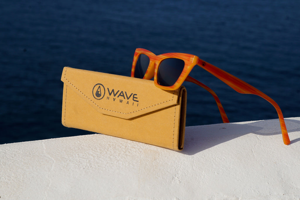 Orangene Fischernetz Sonnenbrille mit Cellulose-Brillencase auf einer weißen Mauer mit blauem Wasser im Hintergrund.