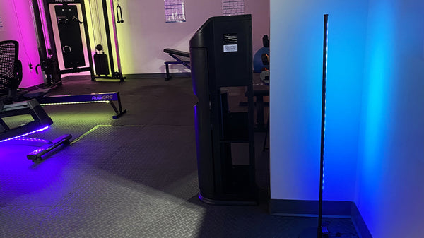 Smart floor lamp in a corner of a gym emitting gradient blue light and other workout machines in the background.