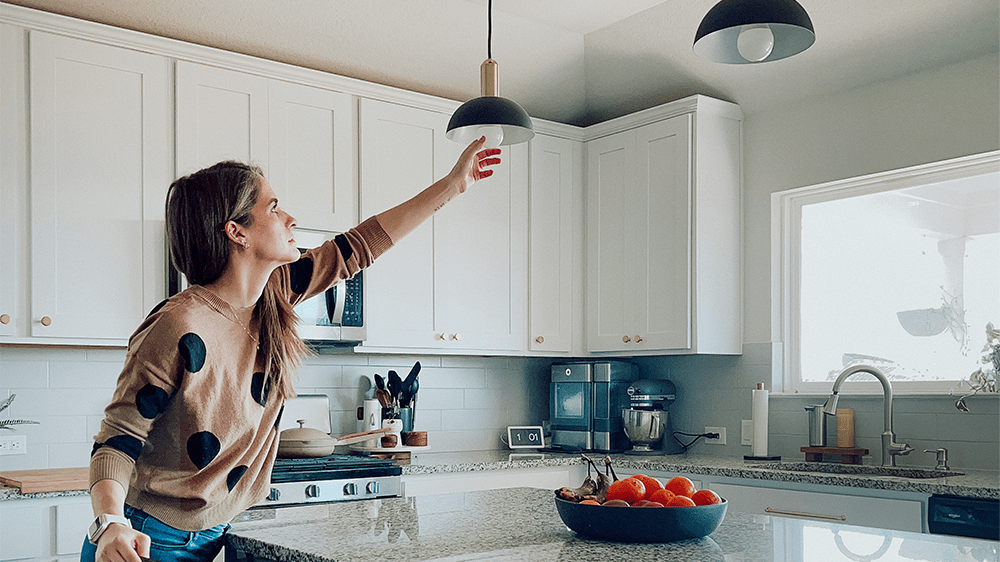 woman in kitchen replacing incandescent bulbs with LED light bulbs in a hanging pendant fixture hanging over kitchen island.