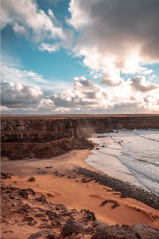 This is a beautiful sundown view of a beach spot called el cottilo, Fuerteventura. You can see the sun, ocean, cliffs, and the orange beach sand.