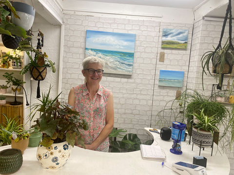 Smiling woman with short hair and glasses standing behind counter in plant shop. There are paintings on the wall behind her.