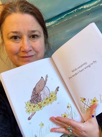 Selfie of Jeanne-Louise holding book open at illustration with Hedgie leaping in daffodil tutu