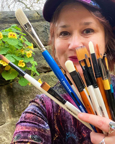 Selfie of Jeanne-Louise a woman wearing a velvet hat and dress. She is holding up a handful of new assorted paintbrushes.