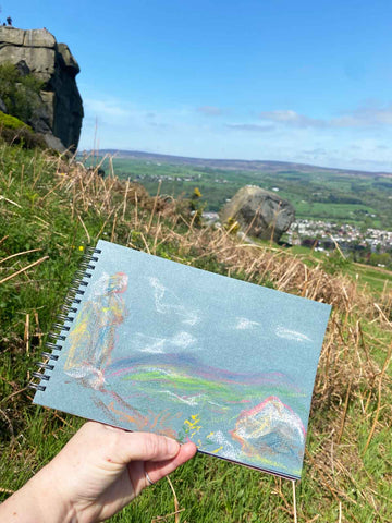 Sketchbook held in front of large stones and outdoor view
