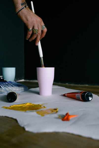 A hand dipping a paintbrush into a cup on the floor.