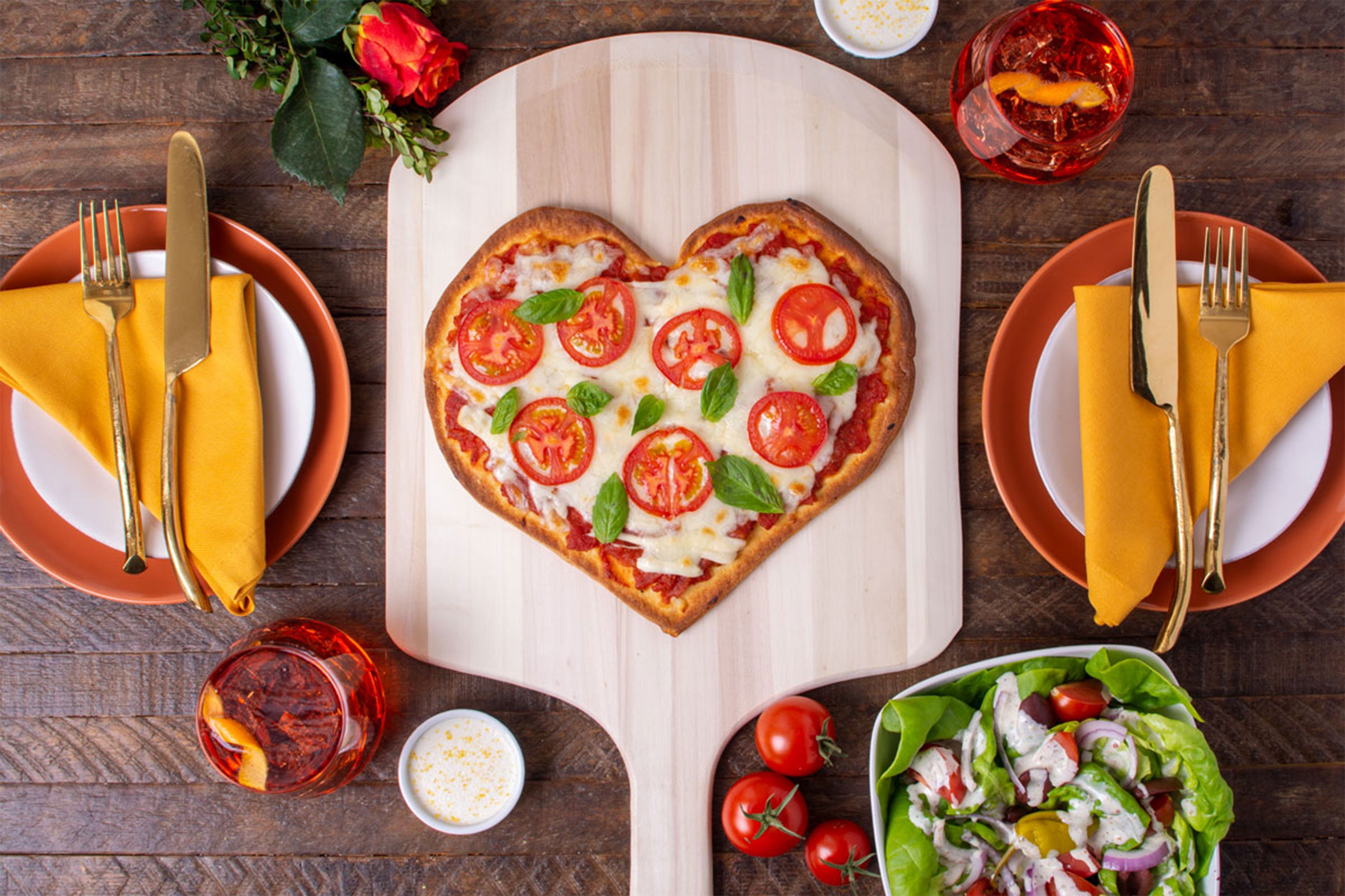 Heart shaped pizza on a pizza board with place servings, drinks, and a salad nearby