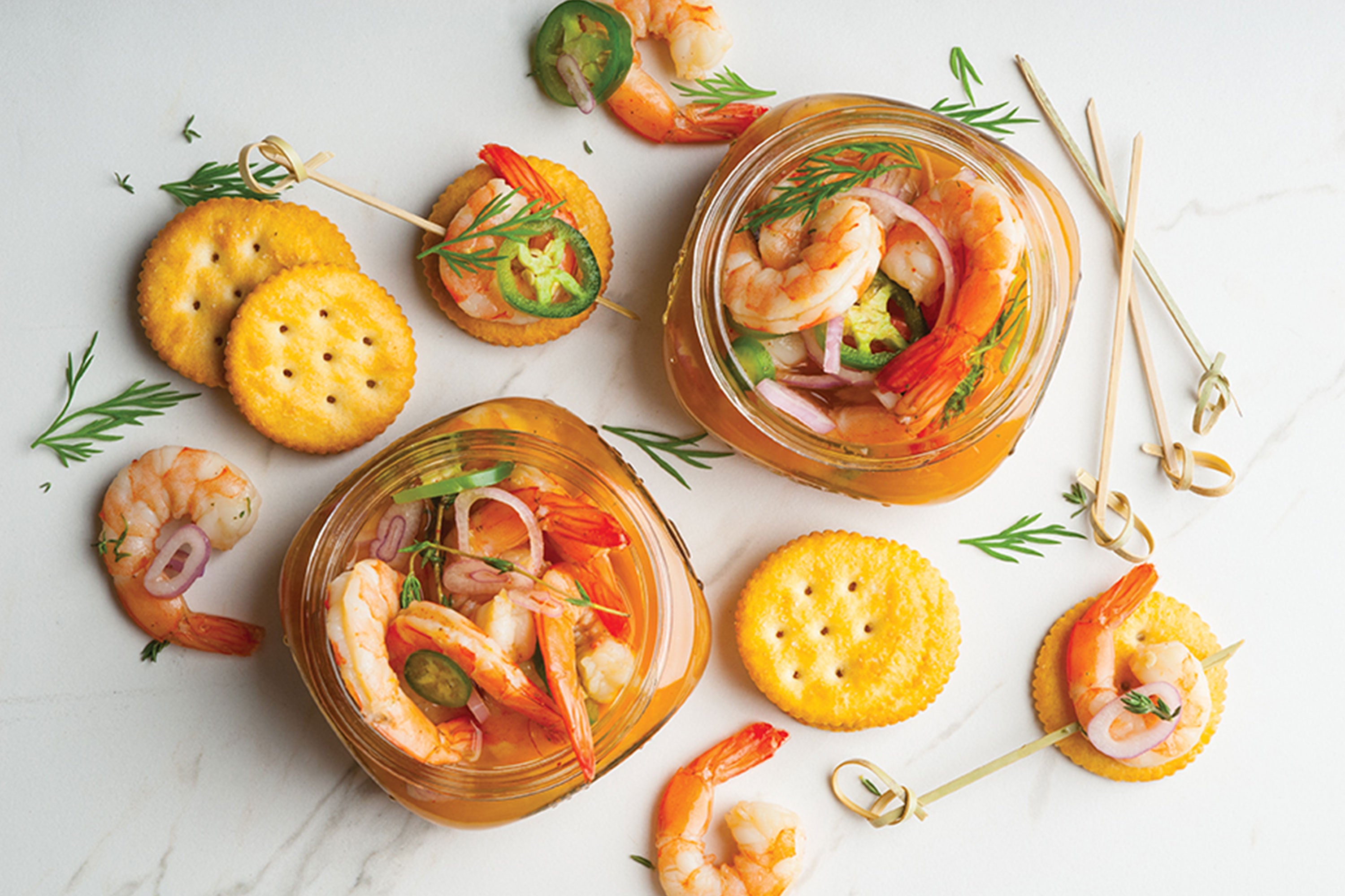 Two jars of pickled shrimp with crackers, small skewers, and pickled vegetables