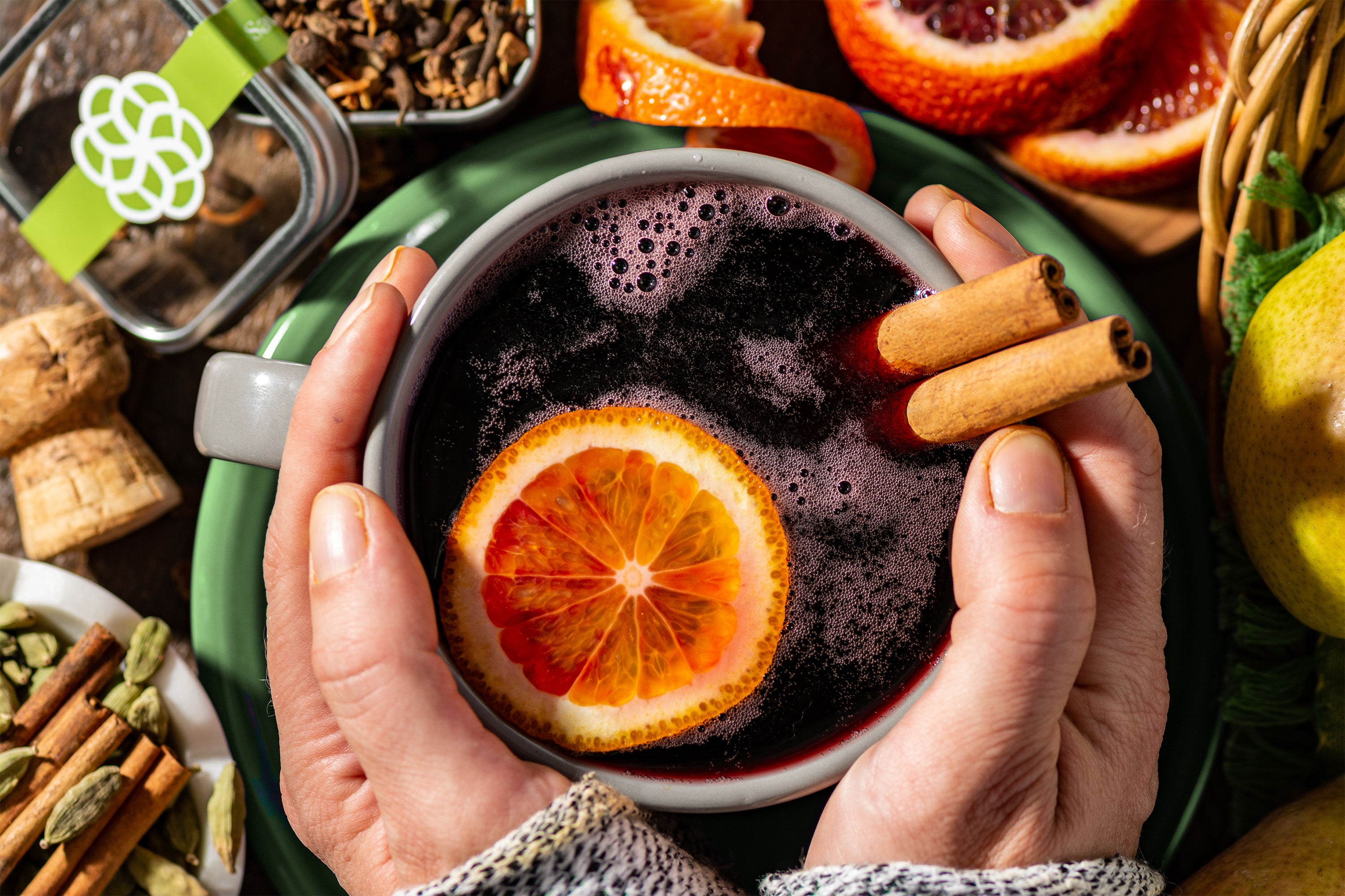 Two hands holding a mug of mulled wine with an orange slice and cinnamon sticks. Assorted ingredients are in the background