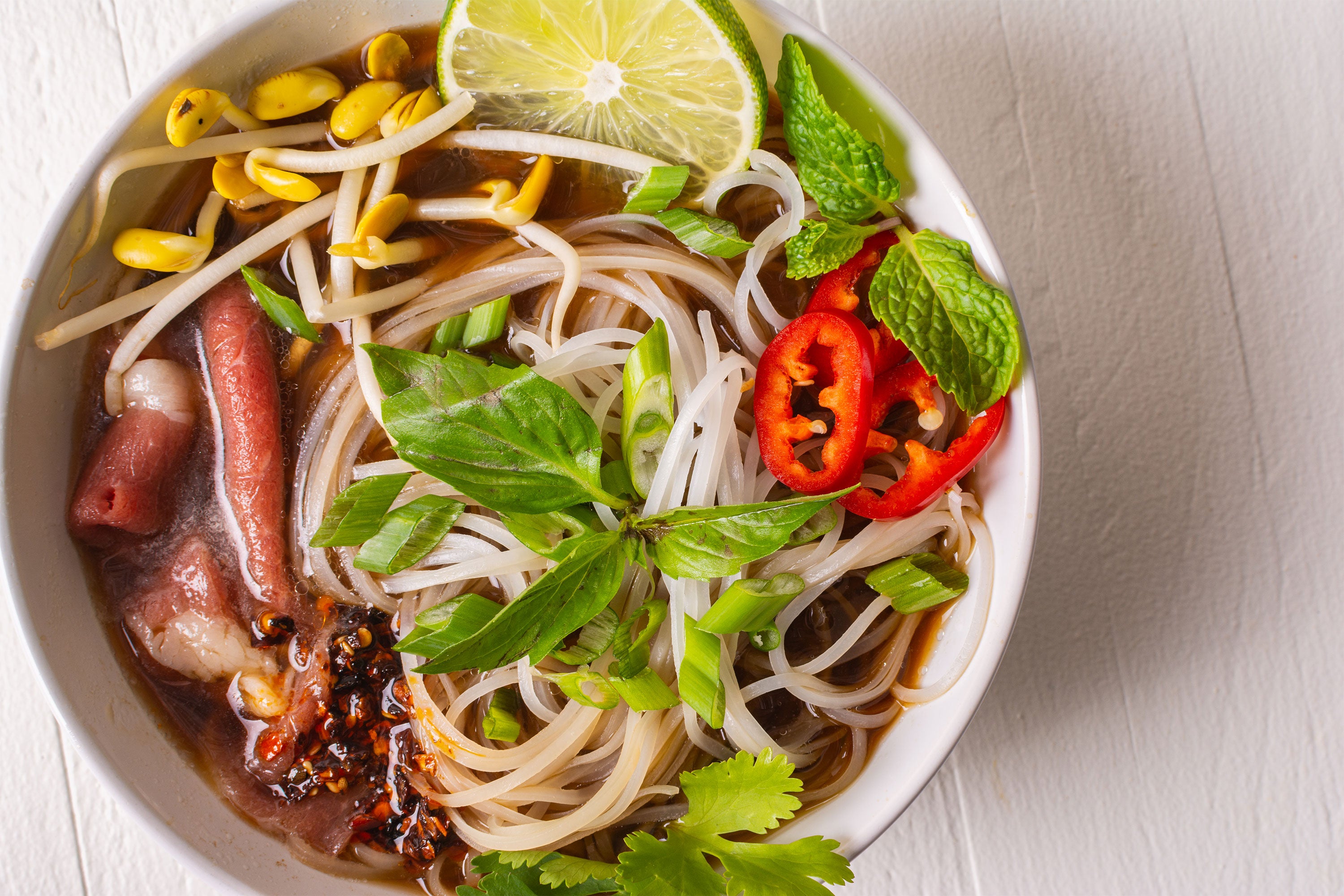 Close up on a bowl of pho topped with herbs, chiles, bean sprouts, and lime slice
