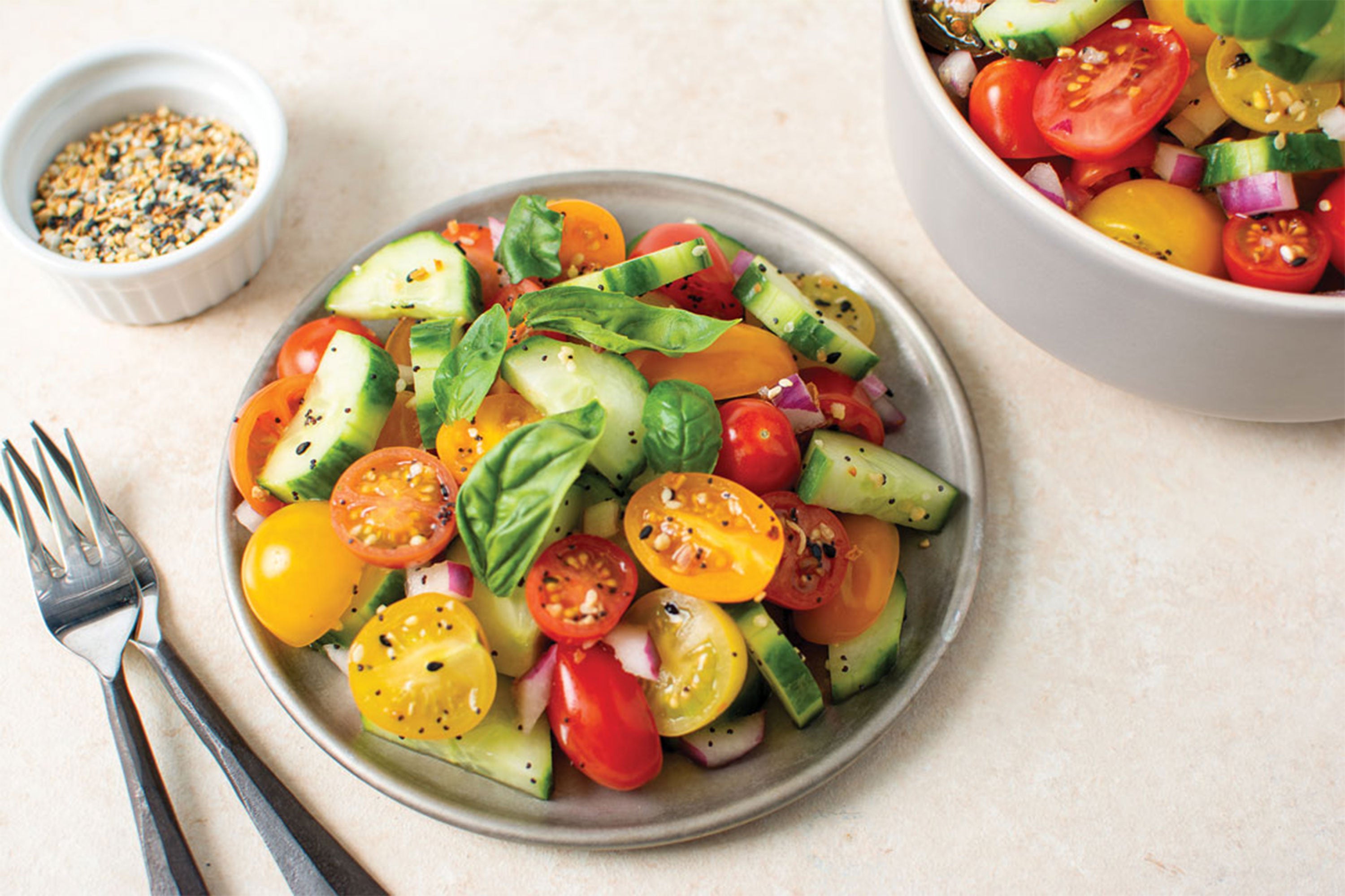 Salad with tomato and cucumber with small bowl of everything bagel seasoning above and silverware to the left