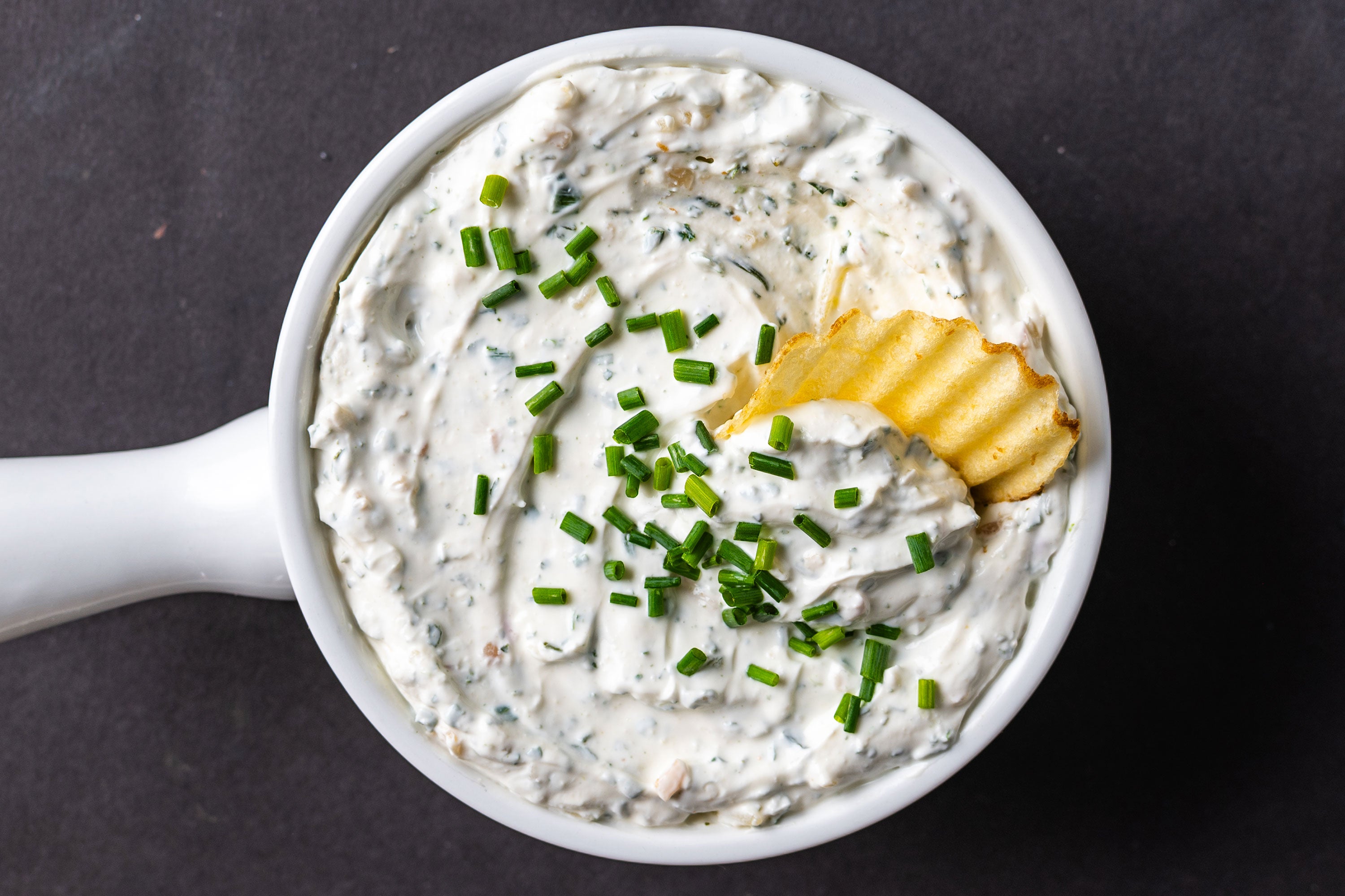 Overhead view of a bowl of onion dip with chives on top and a chip dipped into it