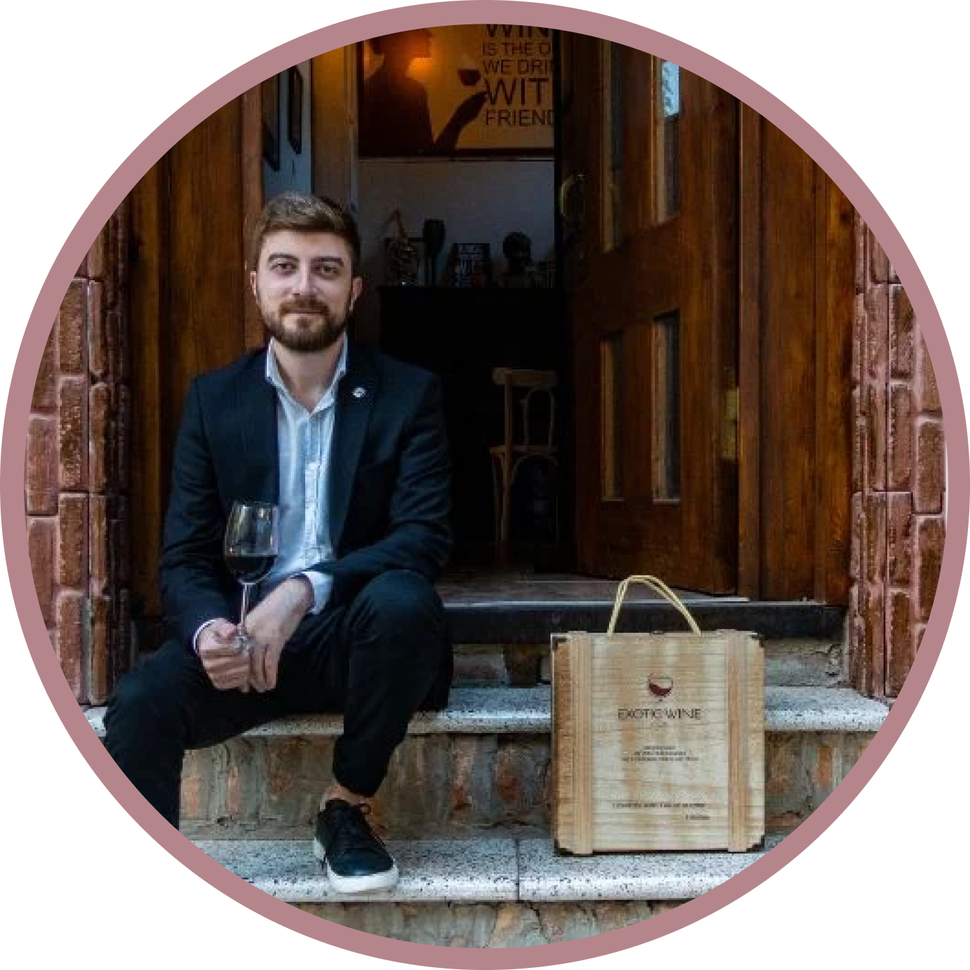 Georgian sommelier Tazo Tamazashvili sitting on stairs, smiling and holding a glass of wine next to a box from Exotic Wine Club.