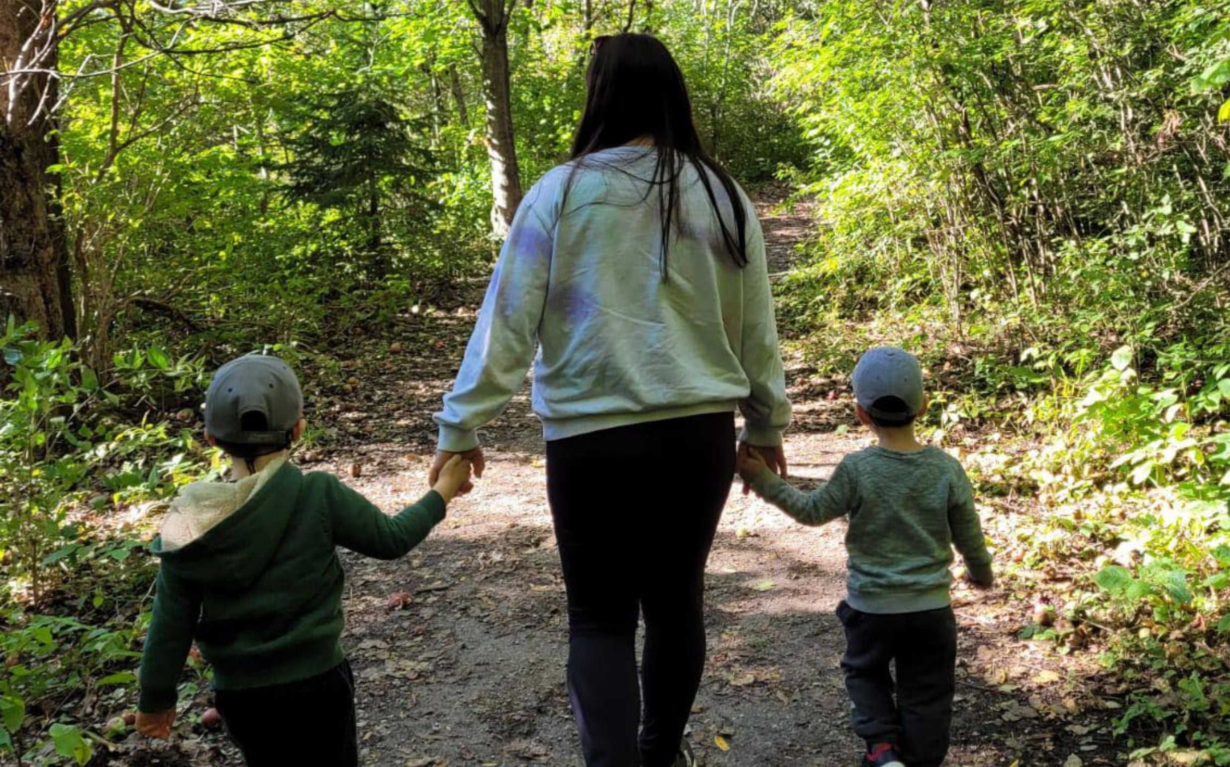 Mom holds hands of two kids facing away from camera hiking into the woods.