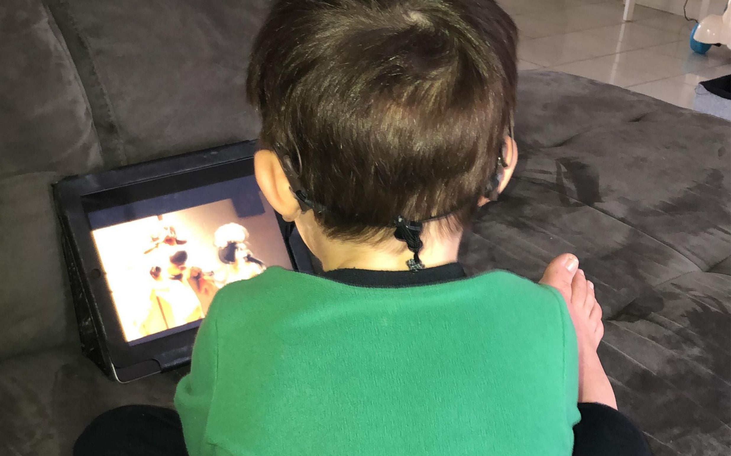 Little boy sits in a green t-shirt playing on a tablet with 2 hearing devices.