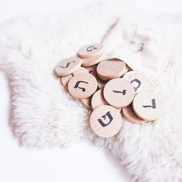 On a white rug a cotton drawstring bag spills out wooden discs marked with letters of the Hebrew alphabet from the Clover + Birch Hebrew Alphabet Hanukkah Gift Set.
